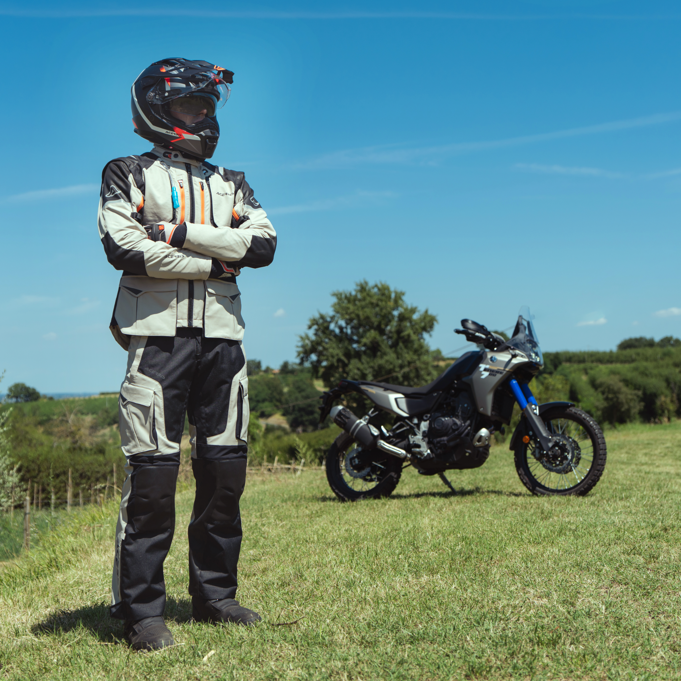 Person in motorcycle gear standing next to a motorcycle in a grassy field with trees in the background