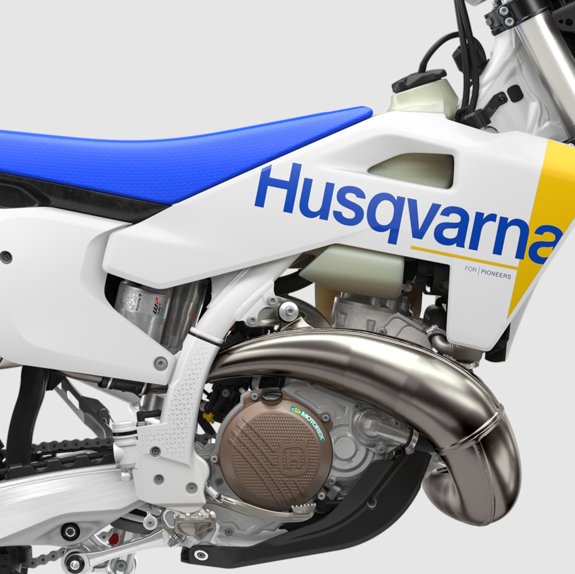 Close-up of a Husqvarna dirt bike showing the white bodywork with blue and yellow graphics, a blue seat, and a polished silver exhaust pipe against a light gray background.