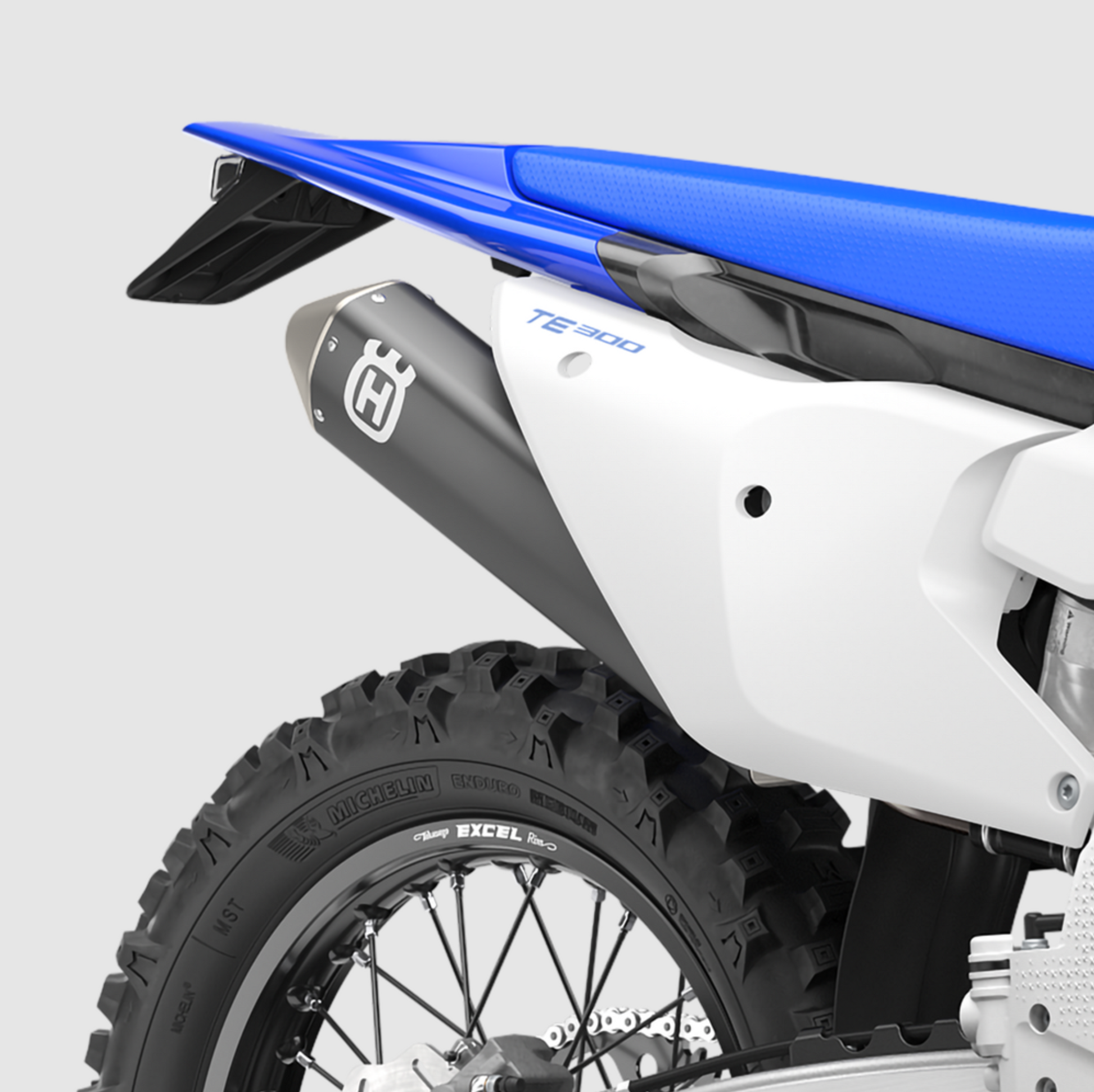 Close-up of a white and blue motorcycle rear section with a black Husqvarna exhaust muffler, rear tire, and blue seat on a light gray studio background.