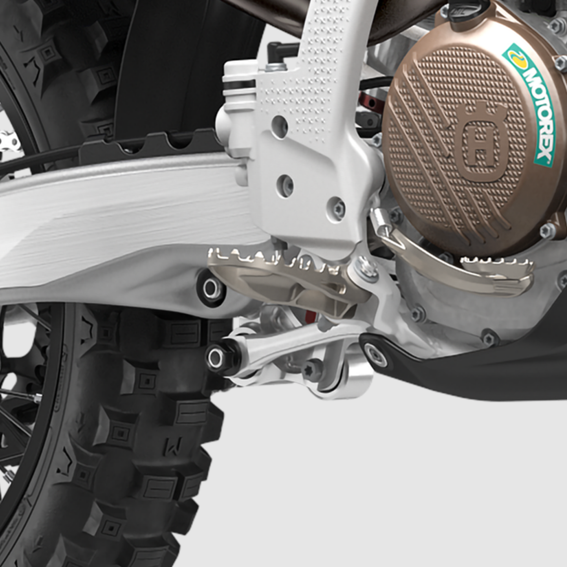 Close-up of a motorcycle's rear suspension and swingarm area, showing a silver swingarm, rear shock, and a bronze-colored engine cover with a visible Motox logo.