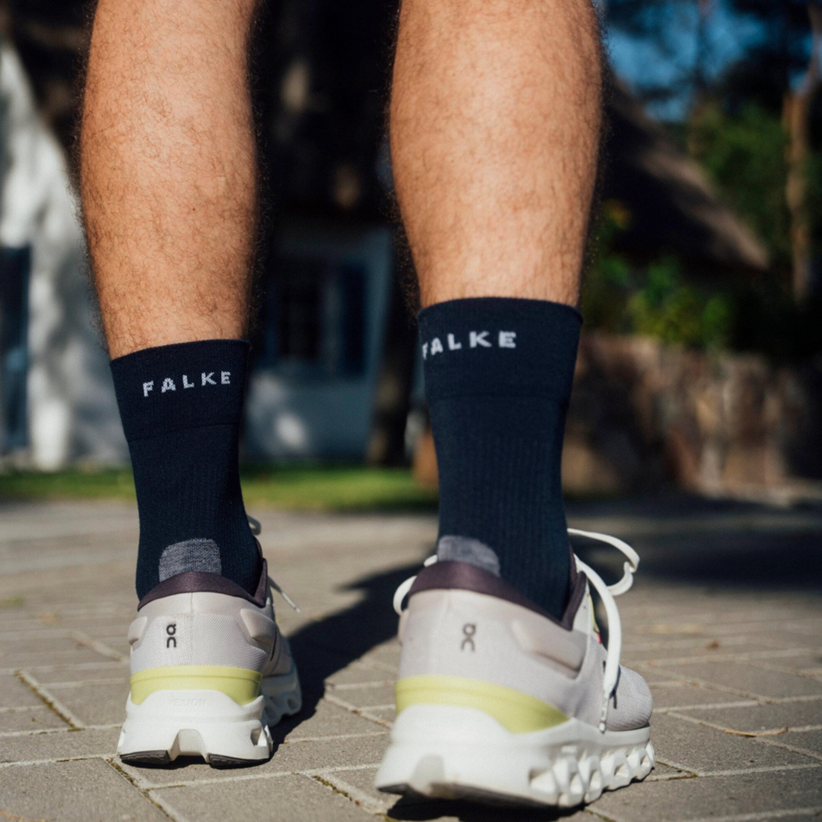 A person standing outdoors on a paved surface, shown from the back, wearing dark navy crew socks with the brand name 'FALKE' in white text and white athletic sneakers.