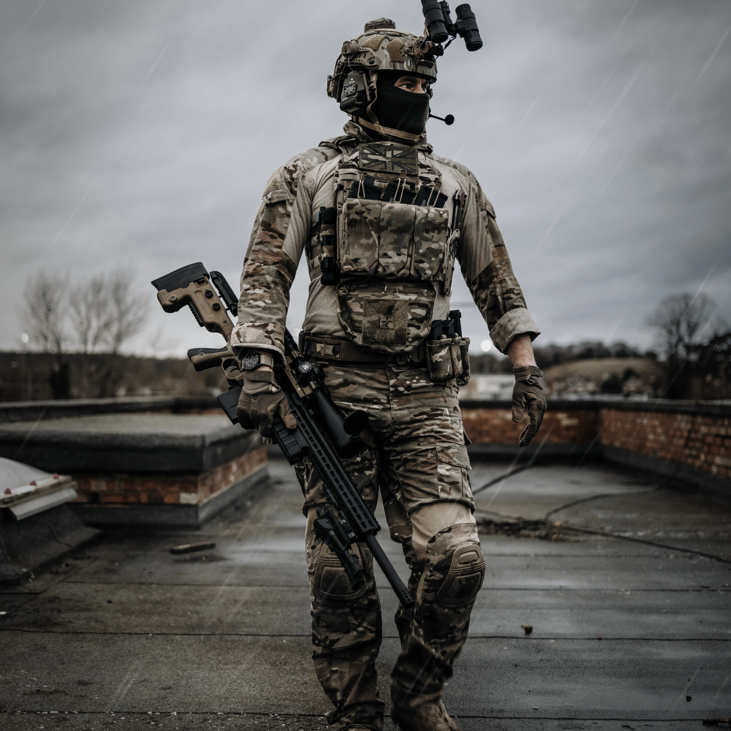 A soldier in full camouflage tactical gear walks across a wet rooftop under a dark, overcast sky, carrying a rifle and wearing a helmet-mounted night vision device.