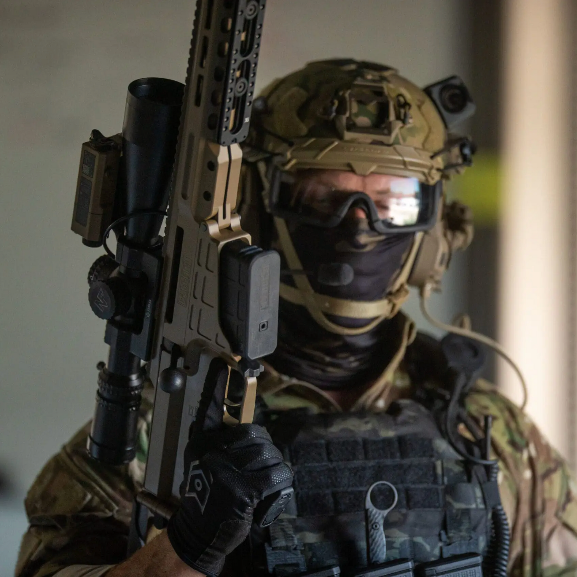 A person in full tactical gear, including a helmet, goggles, face covering, vest, and gloves, holding a tan rifle with a mounted scope. The background is a softly blurred indoor setting.