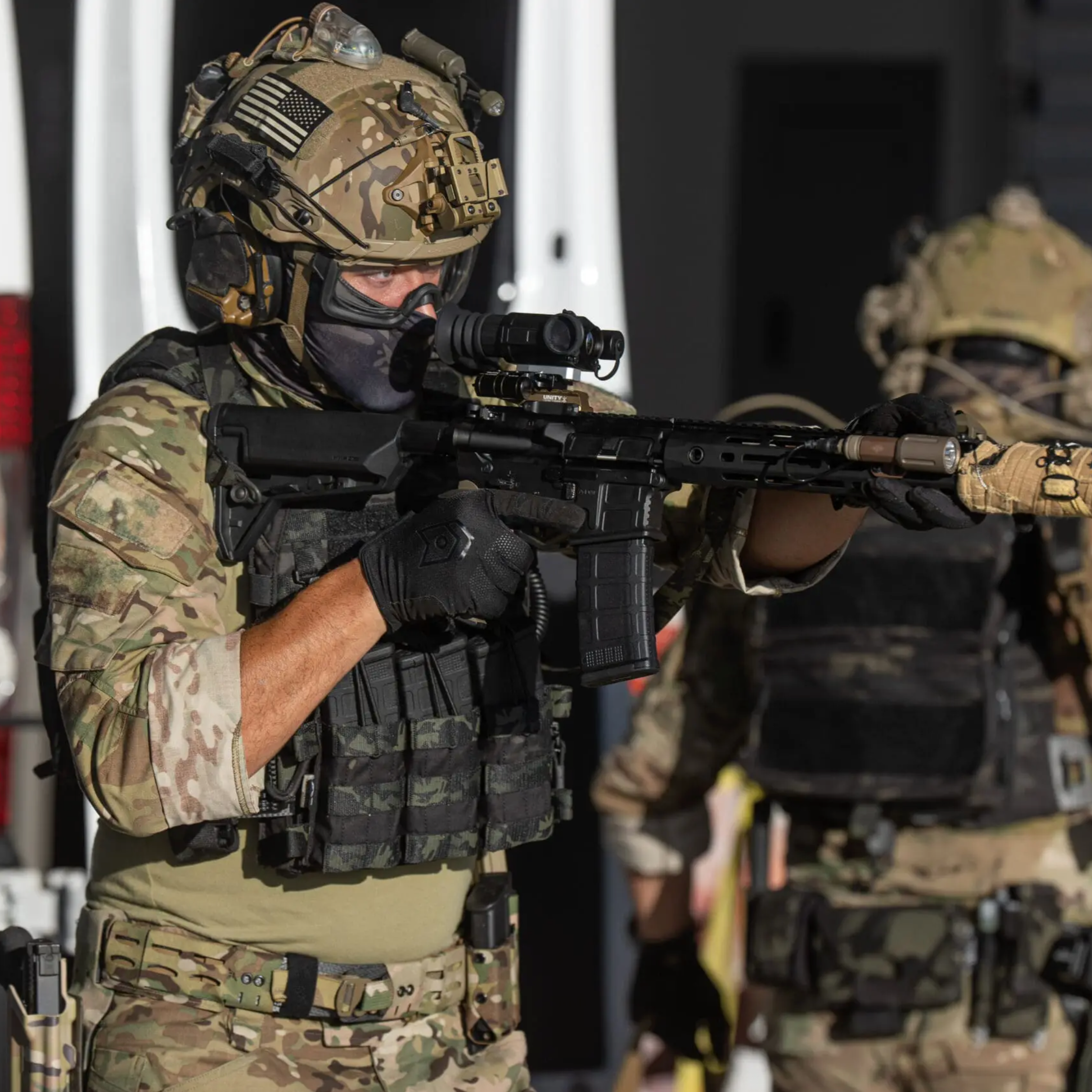 Two tactical soldiers in camouflage uniforms and helmets aim a rifle and another firearm while standing in front of a white van and dark building entrance.