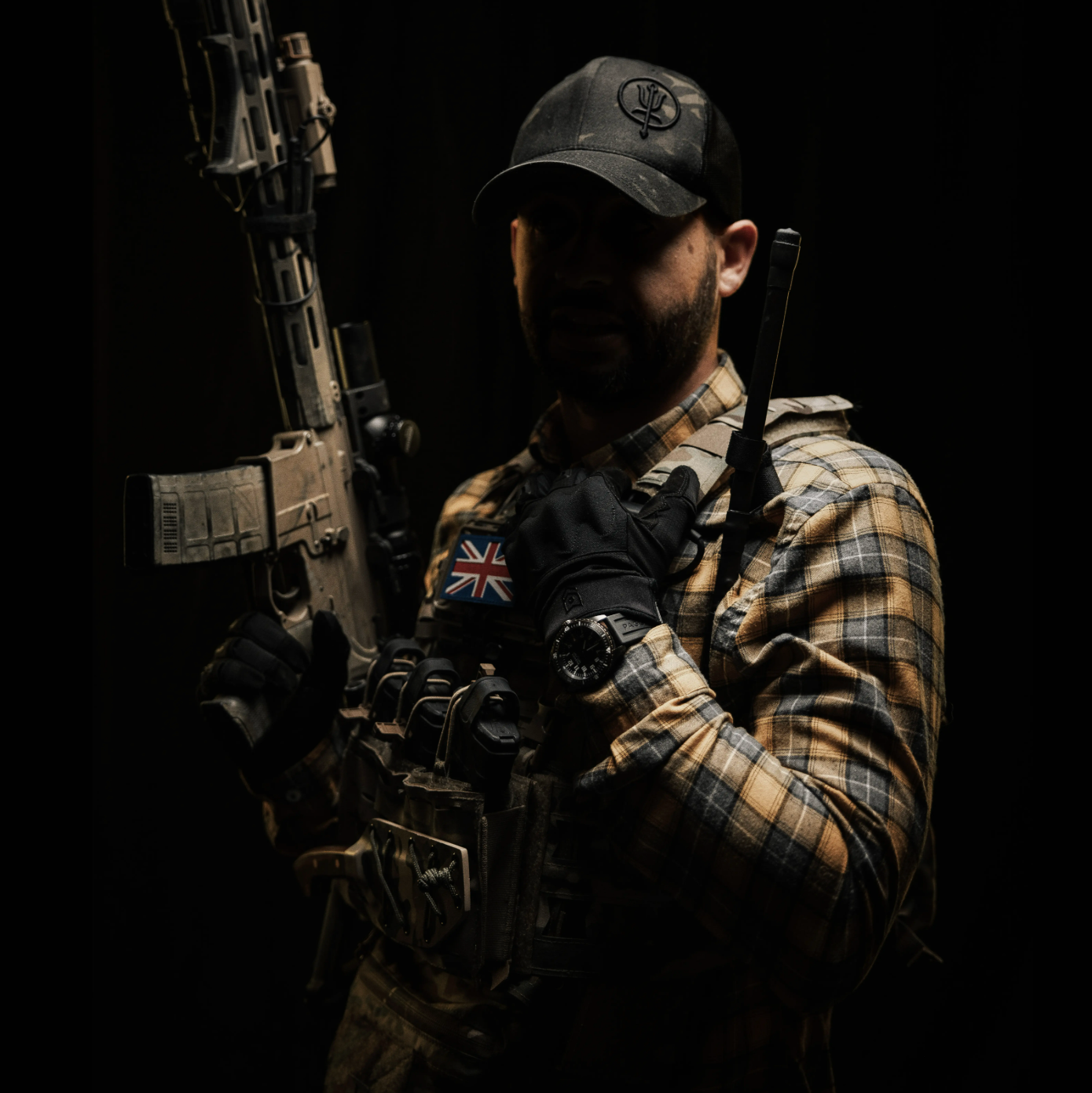 A man in tactical gear, including a plaid shirt, black cap, and gloves, holding a rifle against a solid black background.